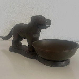 Könnte beinhalten: Eine dunkelbraune Gussmetallfigur eines Hundes, der auf einem Sockel steht, welcher mit einer runden Schale verbunden ist. Der Hund ist im Seitenprofil mit leicht nach vorne gedrehtem Kopf dargestellt und hat eine texturierte Oberfläche.