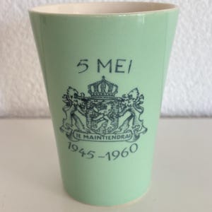 Peut inclure: Un gobelet en céramique vert clair avec un motif imprimé bleu foncé. Le texte indique 5 MEI et 1945-1960, centré au-dessus et en dessous d'un blason détaillé avec une couronne, deux lions et la devise JE MAINTIENDRAI sur une bannière.