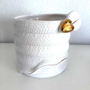 Puede incluir: Taza de cerámica blanca con un diseño texturizado y apilado. El asa presenta un acento de corazón dorado. El borde y la base tienen un ribete dorado. La taza está sobre una superficie blanca.