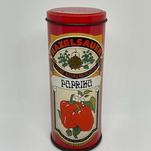 Op de afbeelding: Vintage rode metalen kruidenblik met een deksel. De tekst "HAZELSAUER" staat in een cirkelvormig ontwerp, met daaronder "FIJNE KEUKENKRUIDEN". Een label toont "PAPRIKA" en illustraties van rode paprika's.