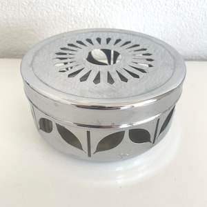 Può includere: Uno scaldavivande rotondo in metallo color argento con un coperchio perforato e ritagli a forma di foglia sui lati. Lo scaldavivande è progettato per contenere un piccolo piatto o pentola e ha una superficie lucida e riflettente.