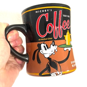 Peut inclure: Mug à café noir avec un motif marron et noir représentant Dingo tenant un plateau avec une cafetière. La tasse porte les mots "Mickey's Coffee" et "Really Swell". La tasse a une anse noire.