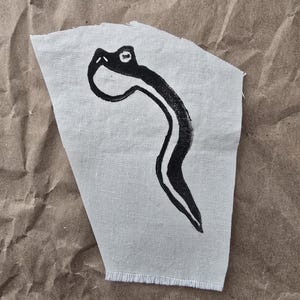 Op de afbeelding: Een stuk lichtgrijze stof met een zwart-witte slangenillustratie. De slang is gestileerd met een gebogen lichaam en een eenvoudige kop. De stof heeft een rafelige rand.