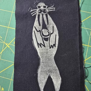 Może przedstawiać: Ciemnoniebieska naszywka z tkaniny z białym nadrukiem linorytu przedstawiającym fokę w okularach przeciwsłonecznych. Foka jest przedstawiona w stylizowanej, abstrakcyjnej formie, z podniesionymi płetwami. Naszywka jest prostokątna.