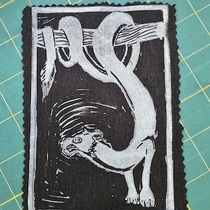 Puede incluir: Un grabado en linóleo en blanco y negro de una criatura colgando de una rama. La criatura tiene un cuerpo largo y serpentino y una cabeza detallada. La impresión está sobre una tela negra con un borde decorativo.