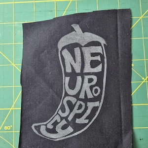 Könnte beinhalten: Ein schwarzer Stoffaufnäher mit einem grauen Chili-Pfeffer-Design und dem Wort "NEUROSPICY" im Inneren. Der Aufnäher ist rechteckig mit rohen Kanten und liegt auf einer grünen Schneidematte.