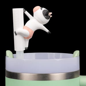 May include: A novelty tumbler with a pale green body, a silver band, and a white and lilac rim. A white and black cartoon cow figure with a pink nappy is attached to a white straw. The cow is posed as if it is pole dancing.