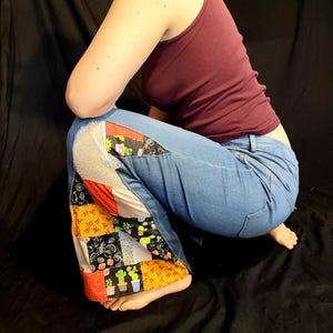 Könnte beinhalten: Hellblaue Jeans mit Patchwork-Einsätzen an den Seiten. Der Patchwork-Stoff weist verschiedene Muster auf, darunter Kakteen, florale Designs und geometrische Formen. Die Person trägt ein weinrotes Tanktop.