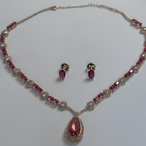 May include: A gold-toned necklace and matching earrings set. The necklace features pink and clear gemstones, with a teardrop-shaped pendant. The earrings have a single pink gemstone. The jewelry is displayed on a white surface.