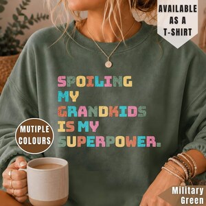 May include: A military green crewneck sweatshirt featuring the multi-coloured text SPOILING MY GRANDKIDS IS MY SUPERPOWER. The garment includes a text overlay stating AVAILABLE AS A T-SHIRT and a circular graphic noting MULTIPLE COLOURS.