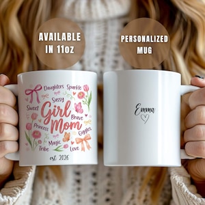 May include: A white ceramic mug featuring a colorful graphic with pink flowers, butterflies, bows, and the text Girl Mom, Daughters Sparkle, Sweet, Sassy, Brave, Princess, Giggles, Magic, Tribe, Love, and est. 2026. The other side displays the name Emma and a heart.
