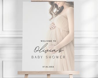 May include: A baby shower sign on a wooden easel. The sign features the text "Welcome to Olivia's Baby Shower" in elegant script. A soft-focus image of a pregnant person in a flowing gown is on the right side of the sign.