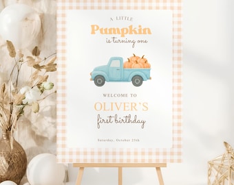 May include: A sign for a first birthday party. The sign features a light blue truck loaded with pumpkins, with the text "A Little Pumpkin is turning one" and "Welcome to Oliver's first birthday". The sign is on a wooden easel.