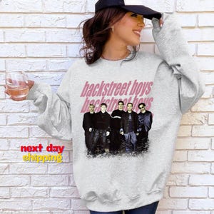May include: A light gray sweatshirt featuring the text "backstreet boys" in pink, with a faded effect. Below the text is a photo of the Backstreet Boys band members. The sweatshirt has long sleeves.