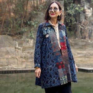 May include: A long, patchwork-style jacket in shades of blue, red, and beige. The jacket features a button-up front and a variety of patterned fabric panels. The model is wearing the jacket over a light-colored shirt and black pants.