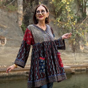 Puede incluir: Una túnica de manga larga con diseño de patchwork. La túnica presenta un cuello en V y una mezcla de patrones, incluyendo secciones de paisley y rayas. La paleta de colores incluye tonos rojos, azules y marrones. La prenda tiene dos pequeños bolsillos y es usada por una persona.