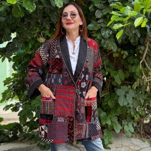 Può includere: Giacca patchwork multicolore con una varietà di motivi, tra cui disegni floreali e geometrici. La giacca ha ampie tasche e un colletto nero. È indossata sopra una camicia bianca e jeans blu. La giacca è nei toni del rosso, nero e blu.