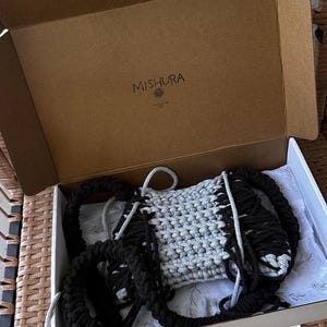 May include: A handmade macrame bag in a cardboard box. The bag features a gray and black woven design with thick black straps. The box is open, revealing the bag inside, with the brand name "MISHURA" printed on the inside of the box.