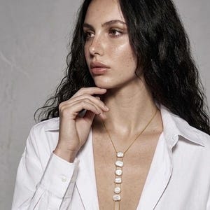 May include: A gold chain necklace featuring five vertically aligned, irregular white baroque pearls. The necklace is worn with a crisp white button-down shirt. The design includes a delicate gold chain that extends downward from the pearls.