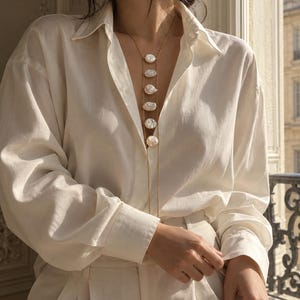 May include: A long gold chain necklace featuring six vertically aligned, irregular white baroque pearls. The necklace is worn over a crisp, long-sleeved white button-down shirt paired with matching white trousers, creating a monochromatic ensemble.