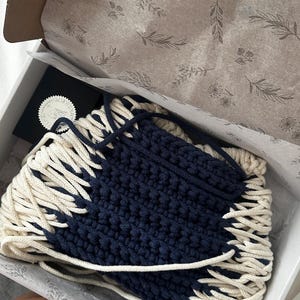 May include: A two-tone crochet bag featuring a dark navy blue center panel and cream-colored side panels with thick, braided cord detailing. The bag is nestled inside a white cardboard box lined with tissue paper printed with botanical illustrations.