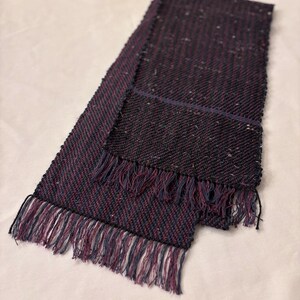 May include: A woven scarf in shades of dark purple, navy, and red with a fringed edge. The scarf is rectangular and appears to be made of a textured fabric. The fringe is a mix of navy and red threads.