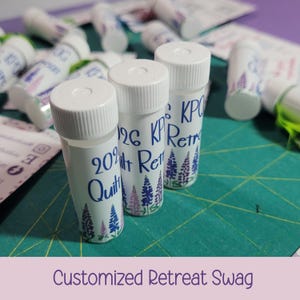 May include: Several small, white cylindrical containers with white lids are arranged on a green surface. Each container has a label with blue text and a floral design in purple and pink. The text includes the words "Retreat" and "Quilt".