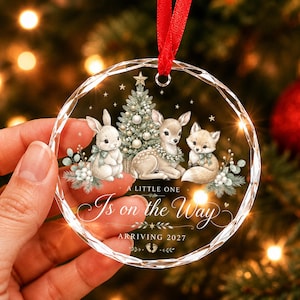 Puede incluir: Un adorno navideño redondo, transparente y facetado de acrílico con una ilustración de un conejo, un cervatillo y un zorro junto a un árbol. El texto dice A LITTLE ONE Is on the Way ARRIVING 2027, con una cinta roja para colgar.