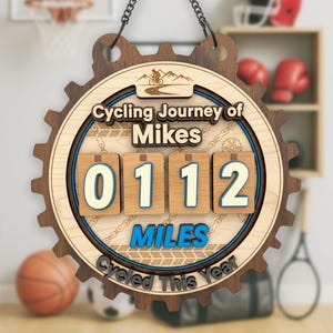 May include: Wooden cycling journey sign with the text "Cycling Journey of Mikes" and "0112 Miles Cycled This Year." The sign is shaped like a gear and features a cyclist graphic and chain design.