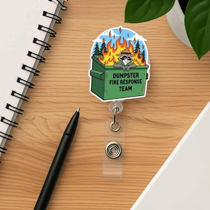 Puede incluir: Un portatarjetas con un mapache de dibujos animados con un sombrero de guardabosques, asomándose de un contenedor de basura verde en llamas. El texto "DUMPSTER FIRE RESPONSE TEAM" está en el contenedor.