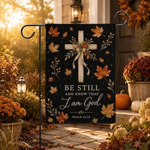 May include: A black garden flag featuring a central wooden cross adorned with autumn leaves and a bow. The text reads: BE STILL AND KNOW THAT I am God. PSALM 46:10. The design is surrounded by scattered orange, yellow, and brown maple leaves.