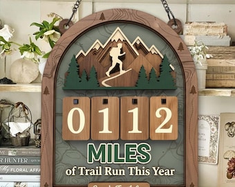 Può includere: Registro di corsa su sentiero in legno con una scena di montagna e la scritta "0112 MILES of Trail Run This Year". Presenta una sagoma di escursionista, alberi verdi e una targhetta personalizzata.