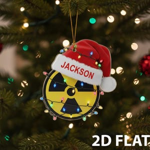 May include: A Christmas ornament featuring a yellow radiation symbol with a red Santa hat that reads "JACKSON". The ornament is decorated with colorful lights and hangs from a gold string. The words "2D FLAT" are at the bottom.