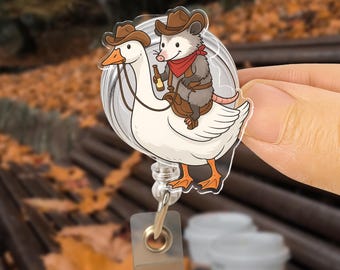Può includere: Un portabadge in acrilico trasparente con un'oca dei cartoni animati e un opossum. L'oca indossa un cappello da cowboy e l'opossum, anch'esso con un cappello da cowboy, lo cavalca, tenendo una bottiglia. Il portabadge ha una clip di plastica trasparente e un cordino retrattile.