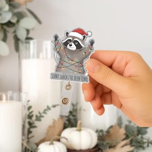 Puede incluir: Un portatarjetas con un mapache de dibujos animados con un gorro de Papá Noel y luces navideñas. El texto "SORRY SANTA I'VE BEEN FERAL" está impreso debajo. El portatarjetas tiene una correa de plástico transparente y un clip metálico.