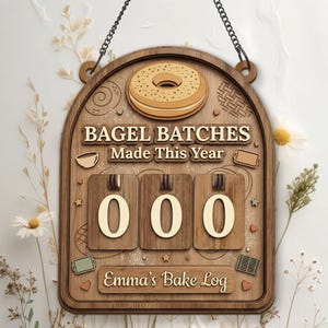 Può includere: Cartello in legno con un'illustrazione di bagel e la scritta "BAGEL BATCHES Made This Year". Presenta tre quadranti numerici impostati su "000" e la scritta "Emma's Bake Log". Include una catena per appendere.
