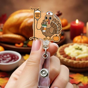 May include: A Thanksgiving-themed badge reel featuring a cartoon turkey wearing a nurse's hat and holding a coffee cup. The reel has a glittery orange background with the text "Pumpkin Spice Energy" on an IV bag. The reel is held in a person's hand.