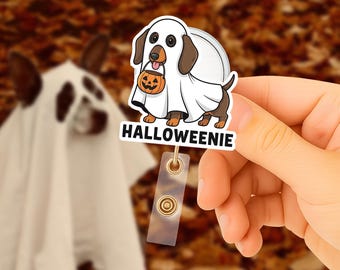Può includere: Un distintivo a tema Halloween con un bassotto vestito da fantasma, che tiene in mano una lanterna a forma di zucca. Il distintivo riporta la scritta "Halloweeniie". Lo sfondo è sfocato, con foglie autunnali.