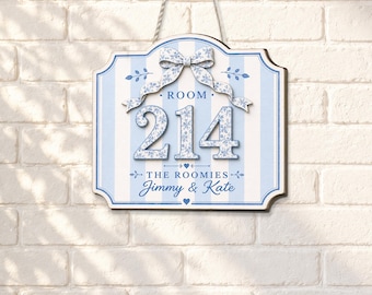 Può includere: Una targa da parete rettangolare con sfondo a righe azzurre e bianche, caratterizzata da un motivo floreale blu sui numeri 214, un fiocco decorativo e il testo ROOM 214, THE ROOMIES e Jimmy & Kate, appesa tramite una corda.