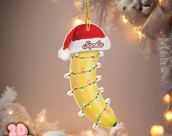 May include: A yellow banana Christmas tree decoration with a red Santa hat that says "Sophie". The banana is adorned with multi-coloured fairy lights and suspended by a gold string. A quirky and festive holiday ornament.