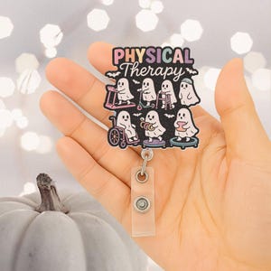 Puede incluir: Un portatarjetas con la frase "PHYSICAL Therapy" e ilustraciones de fantasmas usando equipos de fisioterapia. El portatarjetas se sostiene en una mano y tiene una correa de plástico transparente y un clip de metal.