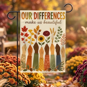 Puede incluir: Una bandera de jardín rectangular con el texto "OUR DIFFERENCES make us beautiful" en marrón y naranja. El diseño muestra ocho manos diversas sosteniendo varios elementos botánicos de otoño, incluyendo hojas de arce, trigo y flores.
