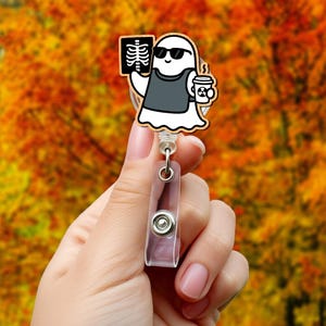 Puede incluir: Un carrete de insignia con forma de fantasma blanco con un diseño de dibujos animados. El fantasma lleva gafas de sol y un chaleco gris, sosteniendo una radiografía y una taza de café. El carrete tiene una correa de plástico transparente y un clip de metal.