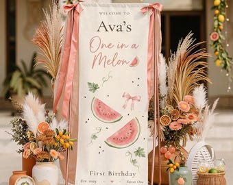Puede incluir: Un letrero decorativo para una celebración de primer cumpleaños. El letrero dice "Welcome to Ava's One in a Melon" con ilustraciones de sandías. El letrero es de color crema con acentos rosados y está sostenido por un marco de metal color cobre.