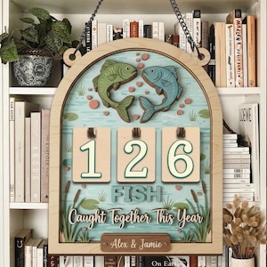 Puede incluir: Letrero de madera con marco de madera clara, con dos peces y el texto "126 FISH Caught Together This Year". El letrero incluye un espacio para nombres y está diseñado con una escena de estanque y una cadena para colgar.