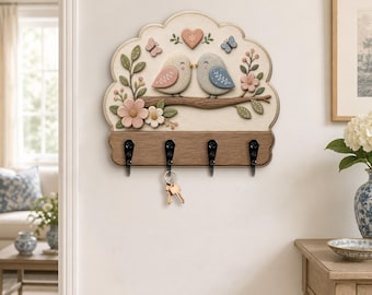Può includere: Un portachiavi da parete con bordo superiore smerlato e un rilievo raffigurante due uccellini su un ramo, circondati da fiori, farfalle e un cuore. La base è color legno con quattro ganci in metallo nero, uno dei quali regge delle chiavi.