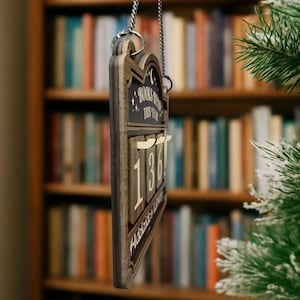 Puede incluir: Contador de libros de madera con el texto "BOOKS READ THIS YEAR" y el n&uacute;mero "136". El contador est&aacute; colgado de una cadena plateada. El fondo muestra una estanter&iacute;a y un &aacute;rbol de Navidad.