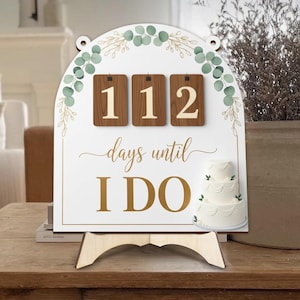 Puede incluir: Un letrero blanco de cuenta regresiva de boda con el texto "112 días para el SÍ, QUIERO". El letrero presenta un pastel de tres niveles y está decorado con detalles verdes y dorados. Se apoya en un caballete de madera.