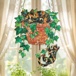 Puede incluir: Un atrapasoles decorativo colgante con gatos. Presenta una maceta de estilo mosaico con un gato durmiendo y un gato trepando. Hojas verdes de hiedra rodean la maceta. Los gatos tienen marcas naranjas, negras y blancas.
