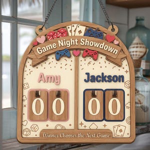 以下が含まれることがあります： 「Game Night Showdown」の文字が入った木製のゲームナイトスコアボード。2人分のスペース、サイコロ、ハート、トランプ、ゲームコントローラーが特徴です。「Amy」と「Jackson」の名前がスコアボックスの上に表示されています。